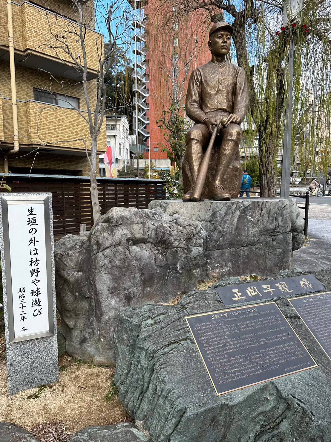 Bronze statue of Masaoka Shiki in baseball uniform holding a bat, with haiku plaque reading ็ๅฃใฎๅคใฏๆฏ้ใ็้ใณ